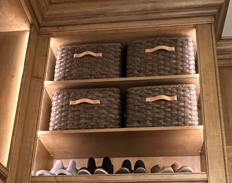 Four dark-stained handwoven baskets with leather handles, two to a shelf, filling the top of a lit closet cabinet